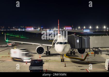 Un avion se tient sur la passerelle d'embarquement la nuit avec un avion en mouvement en arrière-plan Banque D'Images