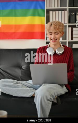 Portrait d'un jeune homme gay asiatique heureux utilise son ordinateur portable tout en se relaxant sur un canapé dans son salon. Banque D'Images