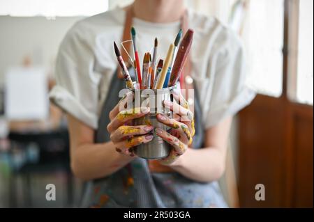 Gros plan d'un jeune artiste masculin dans un tablier tenant un porte-crayon avec différents crayons et pinceaux dans son studio. Banque D'Images