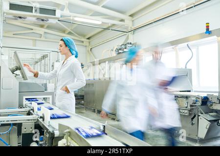 Une femme occupée dans des vêtements stériles en choisissant un programme sur écran tactile tout en faisant fonctionner la machine de fabrication de produits alimentaires emballés Banque D'Images