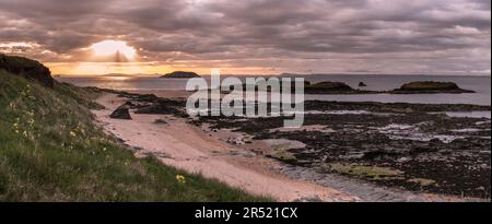 Coucher de soleil et rayons du soleil dans un ciel nuageux ; Craigleith Island dans le Firth of Forth avec Milsey Bay Beach à marée basse et des glissements de bois sauvages en premier plan Banque D'Images