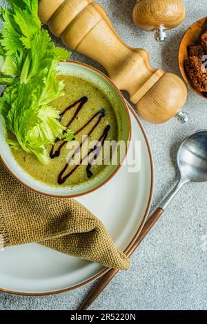 Vue de dessus de la soupe de crème de céleri saine dans un bol avec des branches de céleri sur l'assiette avec une serviette, une cuillère, des bocaux et un bol avec du pain sur fond gris Banque D'Images