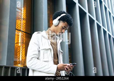 Petit angle de jeune Afro-américain avec casque sans fil et une coiffure afro-américaine qui écoute de la musique tout en parcourant son smartphone dans la rue Banque D'Images
