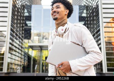 Petit angle de jeune homme afro-américain joyeux avec des écouteurs autour du cou et un ordinateur portable souriant comme il regarde l'autre Banque D'Images