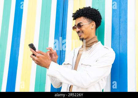 Jeune Afro-américain avec des lunettes de soleil et une coiffure africaine texting avec un téléphone mobile et souriant alors qu'il fixe l'écran contre un mur Banque D'Images