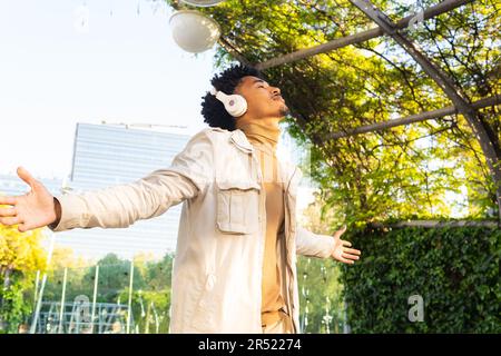 Joyeux jeune Afro-américain avec les yeux fermés à l'écoute de musique avec des écouteurs avec les bras étendus pendant qu'il marche contre les arbres dans le parc Banque D'Images