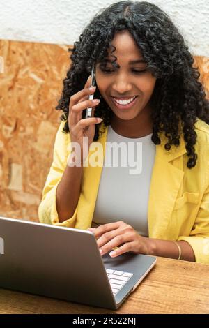 Bonne jeune femme afro-américaine se concentrant sur un bureau avec un ordinateur portable et parlant d'un projet d'entreprise pendant un appel téléphonique Banque D'Images