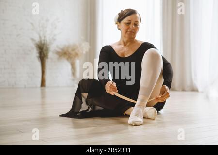 Corps complet de danseuse mature en robe noire assise sur le sol et mettant sur le ballet pointe chaussures tout en préparant pour la répétition en studio Banque D'Images