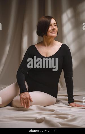Danseuse d'âge moyen décontractée vêtue d'un costume noir et de collants posés sur un tissu froissé et regardant loin dans un studio Banque D'Images