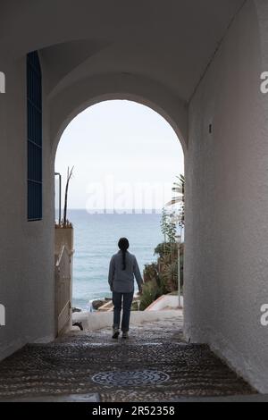 Vue arrière de la femelle anonyme qui sort du bâtiment résidentiel avec des murs blancs et une sortie d'arche située près de la mer calme sous le ciel bleu à Malaga Espagne Banque D'Images