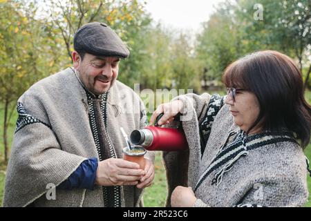 Des amis dans des vêtements chauds se tenant près des buissons verts et versant la boisson chaude des thermos dans la tasse tout en passant du temps ensemble le jour ensoleillé Banque D'Images