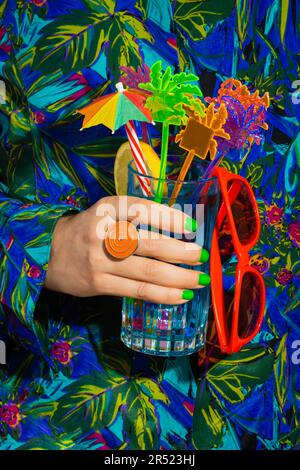 Personne en forme de vêtements colorés tenant un verre à cocktail avec des fleurs d'orange et de menthe, de la paille et des lunettes dans la composition colorée Banque D'Images