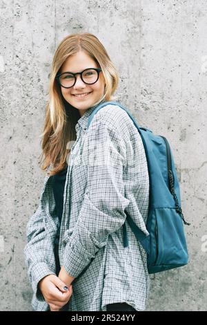 Portrait en plein air d'une jeune fille adolescente portant des lunettes et un sac à dos, posant sur un fond de mur gris Banque D'Images