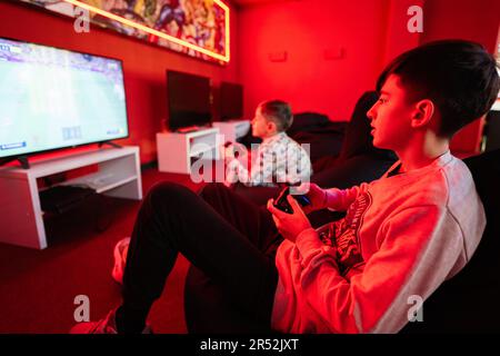 Deux gamers de garçons jouent à la console de jeu vidéo de manette de football dans la salle de jeu rouge. Banque D'Images