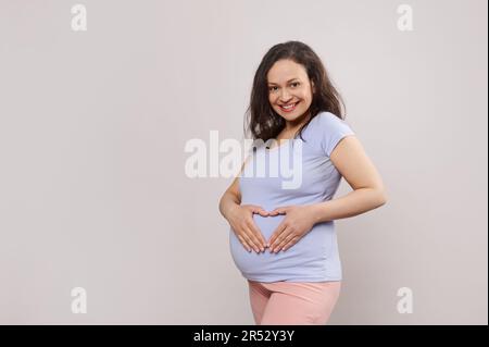 Belle femme enceinte heureuse tenant les mains sur son ventre, formant une forme de coeur, regardant la caméra, isolée sur blanc Banque D'Images