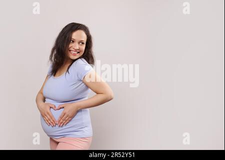 Bonne femme enceinte, femme enceinte souriante en regardant de côté, formant le coeur avec les mains sur le ventre, isolé sur blanc Banque D'Images
