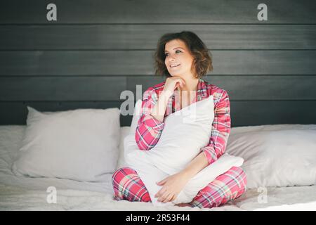 Portrait intérieur de la belle jeune femme se relaxant dans le lit, tenant le coussin, portant rouge confortable pyjama Banque D'Images