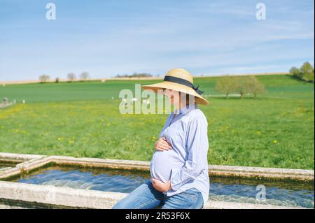 Portrait extérieur de la jeune femme enceinte heureuse appréciant une belle journée à la campagne, assis sur la fontaine d'arrosage, pâturage vert avec des vaches sur le fond Banque D'Images