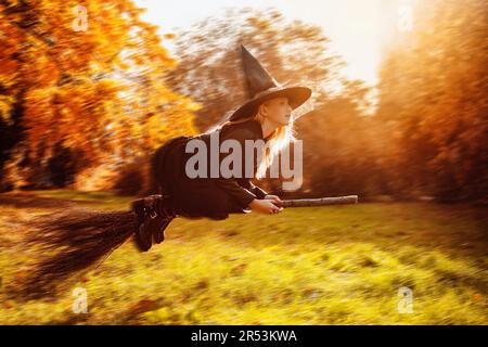 La petite sorcière vole sur un bâton de bois dans la forêt d'automne au coucher du soleil. Banque D'Images