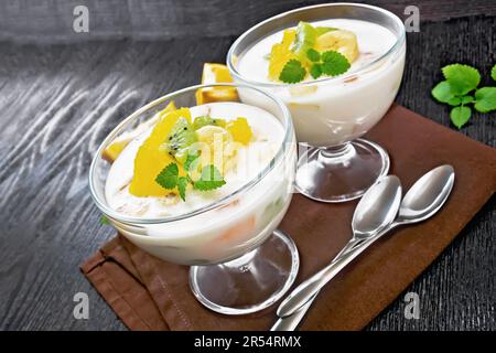 Dessert yaourt aéré avec orange, banane et kiwi dans deux bols en verre, fruits, cuillères et menthe sur une serviette brune sur fond de bois Banque D'Images