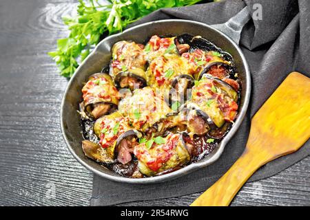 Roulés d'aubergines cuits avec de la viande, des tomates et du fromage dans une casserole, une serviette, du persil et une spatule sur fond de bois Banque D'Images