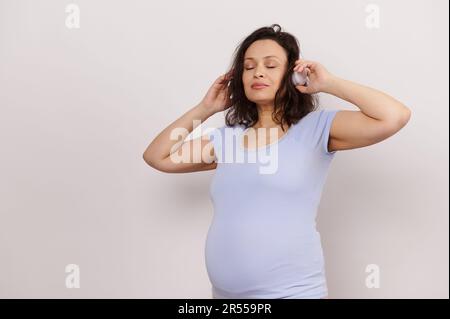 Bonne femme enceinte adulte qui écoute de la musique sur un casque sans fil, se posant avec ses yeux fermés sur fond blanc Banque D'Images