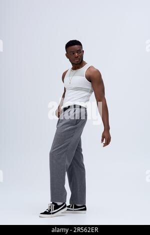 Pantalon et débardeur sur toute la longueur d'un homme afroamerican résistant, posé avec assurance dans une tenue élégante et tendance sur fond gris Banque D'Images