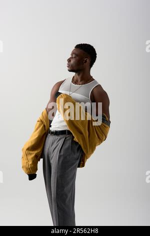 Vue latérale d'un superbe modèle afro-américain en blouson d'aviateur et t-shirt sans manches posé isolé sur une photo grise et contemporaine avec une élégante batterie Banque D'Images