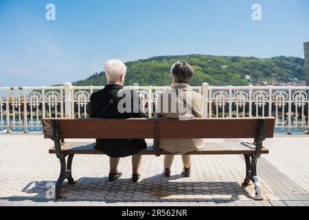 Femmes amies, vue arrière de deux amies seniors assises ensemble sur un banc sur la promenade de San Sebastian - Donostia, Espagne. Banque D'Images