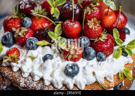 Gâteau frais de saison en couches avec des fruits mûrs printaniers comme des fraises, des myrtilles et des cerises. Placé sur une élégante assiette bleue sur une planche de bois rustique Banque D'Images