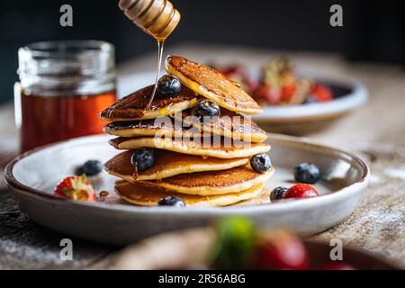 Crêpes américaines aux myrtilles, fraises et cerises avec trempette de miel. Placé sur une assiette de designer rustique, sur une planche rustique en bois. Banque D'Images