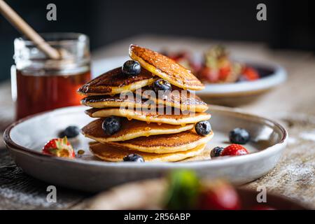 Crêpes américaines aux myrtilles, fraises et cerises avec trempette de miel. Placé sur une assiette de designer rustique, sur une planche rustique en bois. Banque D'Images