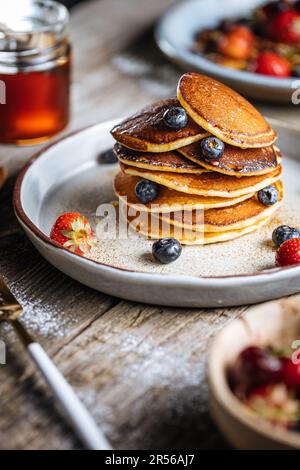 Crêpes américaines aux myrtilles, fraises et cerises avec trempette de miel. Placé sur une assiette de designer rustique, sur une planche rustique en bois. Banque D'Images