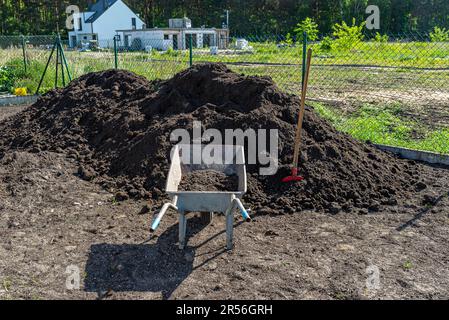 Un tas de terre noire pure se trouvant dans la cour à côté de la clôture, une pelle visible et une brouette vide. Banque D'Images