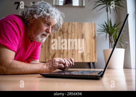 Homme âgé en chemise rose couché sur le sol du salon en tapant sur un ordinateur portable dans une image conceptuelle de l'éducation et de l'apprentissage de nouvelles compétences à la retraite Banque D'Images