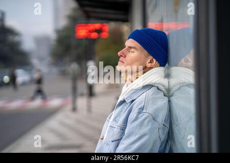 Un petit hipster fatigué assis avec les yeux fermés à l'arrêt de bus Banque D'Images