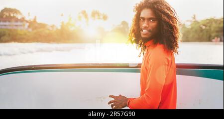 Portrait d'un adolescent noir à poils longs avec planche de surf prête pour le surf avec rétroéclairage au coucher du soleil. Il marche dans les vagues de l'océan Indien. Sports nautiques extrêmes Banque D'Images