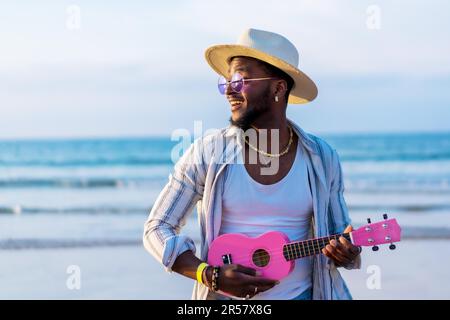 Homme ethnique noir jouant ukulele au bord de la mer en profitant des vacances d'été sur la plage Banque D'Images