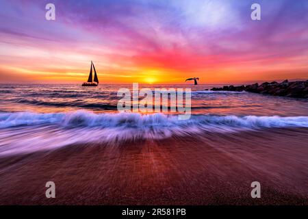 Un beau coucher de soleil sur l'océan avec Une vague se brisant sur la rive et un Voilier en voilier sur Horizon Banque D'Images