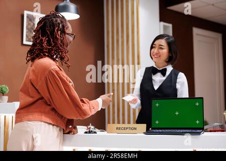 Femme donnant la carte-clé de la chambre au touriste, utilisant un ordinateur portable avec écran d'accueil isolé sur le bureau de réception. Le personnel de l'hôtel travaille à la réception avec un modèle de maquette d'espace publicitaire vierge. Banque D'Images