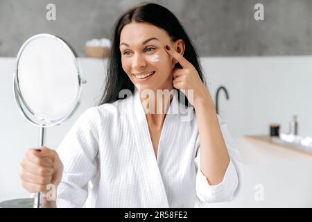 Magnifique femme caucasienne bien entretenue dans un peignoir, tient et regarde un miroir, applique une crème hydratante anti-âge sur son visage, empêche le vieillissement de la peau et sèche. Traitements hydratants quotidiens pour la peau Banque D'Images