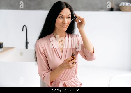 Routine beauté. Hydratation quotidienne de la peau. Jolie femme caucasienne brune, dans un peignoir, applique un sérum hydratant anti-âge avec de l'acide hyaluronique sur son visage, regarde loin, sourit Banque D'Images