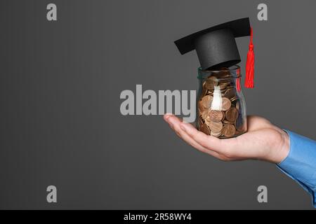 Homme tenant un pot de pièces en verre et un capuchon de graduation sur fond gris foncé, gros plan avec un espace pour le texte. Concept de bourse Banque D'Images