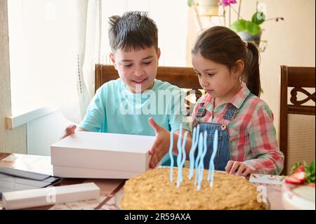 Portrait authentique d'un adorable garçon caucasien ouvrant le cadeau d'anniversaire, assis à table avec un gâteau d'anniversaire. Banque D'Images