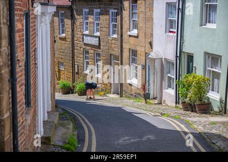 Narrow Street avec un homme en train de désherber les pavés à l'extérieur de sa propriété à Robin Hoods Bay, North Yorkshire, Royaume-Uni Banque D'Images