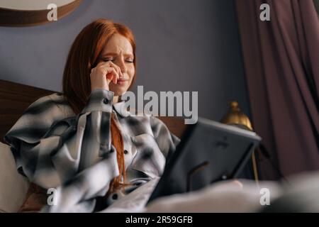 Gros plan d'une jeune femme en deuil, allongé sur un lit, tenant une photo dans le cadre et en pleurant, touchant la photo avec amour, essuyant les larmes avec le mouchoir. Banque D'Images