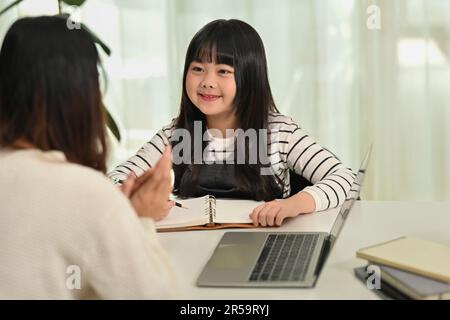 Joyeux petit enfant asiatique assis à un bureau faisant l'affectation de tâche à la maison avec la mère dans le salon Banque D'Images