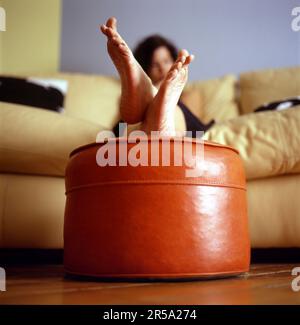 La jeune femme est assise sur un canapé en cuir jaune avec les pieds sur le hassock orange. Banque D'Images
