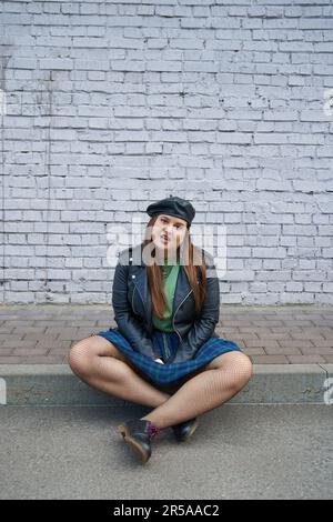 femme chic de grande taille posant dans une veste en cuir, jupe à carreaux avec chaînes, collants résille et chaussures noires tout en étant assise près d'un mur de briques Banque D'Images