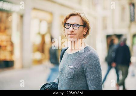 Portrait extérieur d'un bel homme à cheveux rouges portant des lunettes se posant sur la rue de la ville Banque D'Images